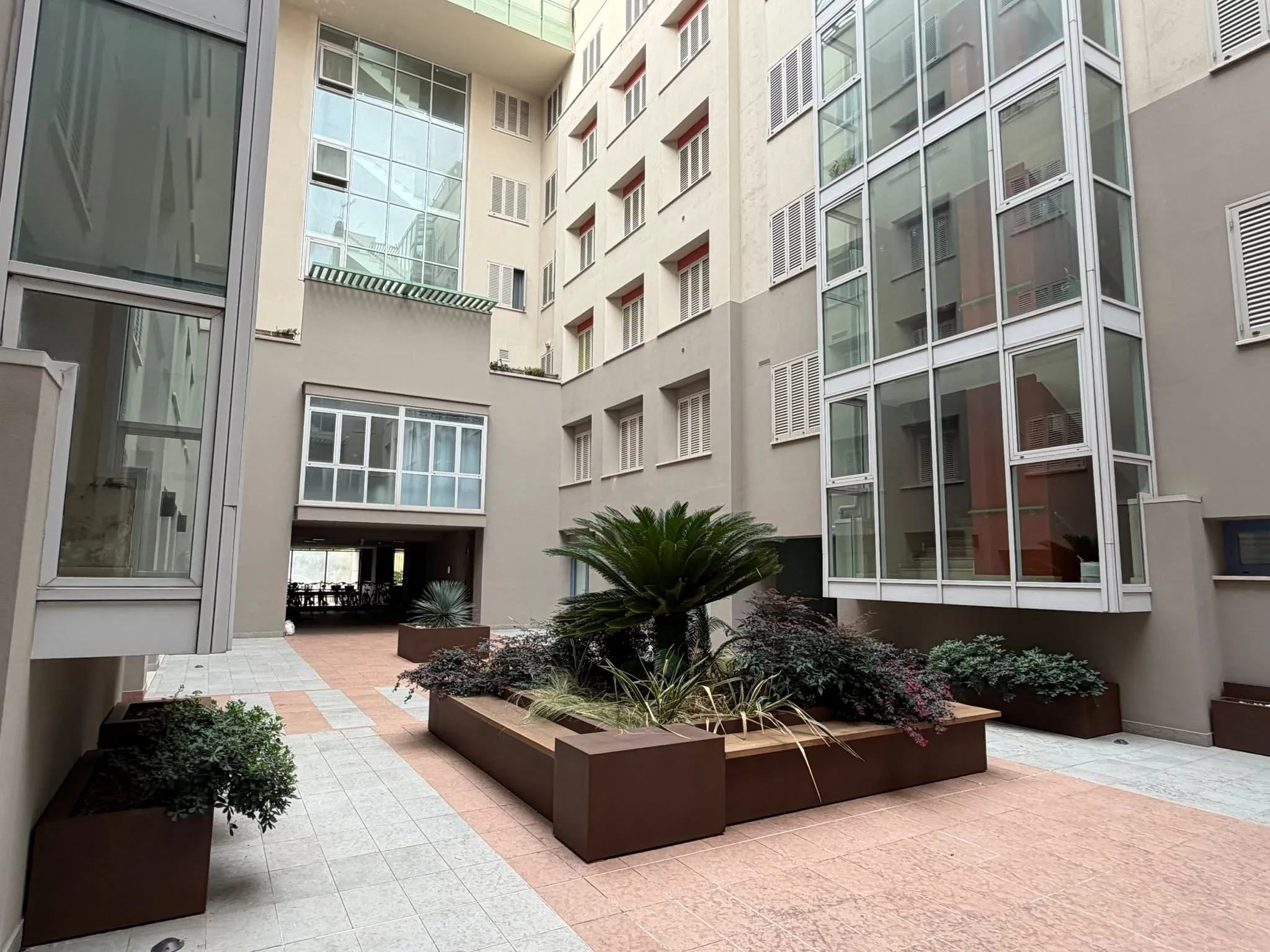 Cortile interno dell'edificio con fioriere centrali, palme e grandi vetrate verticali della scala